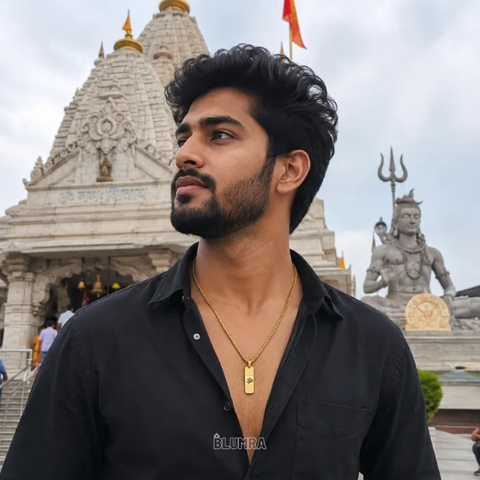 A man wearing a black button-up shirt and a gold pendant necklace stands in front of a white temple with Shiva statues and a cloudy sky.