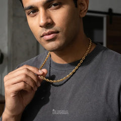 A man wearing a dark gray t-shirt and a gold chain necklace, holding the chain with his hand. The brand name 'BLUHRA' is printed on the chest of the shirt.